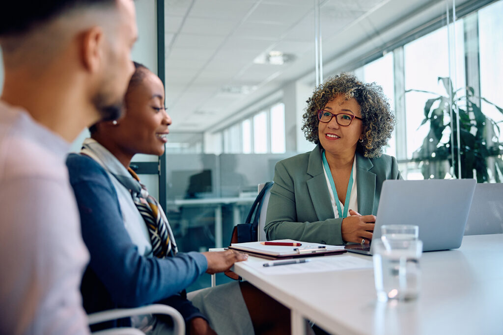 Business meeting around a table in a glass office with two ladies and the side view of a man, clipboard on table.